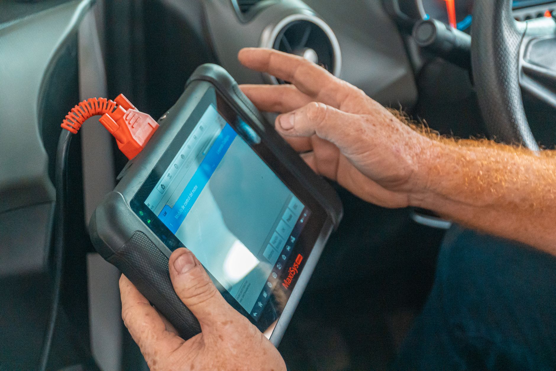 Nahaufnahme eines Diagnoseinterfaces am OBD-II-Anschluss eines Elektroautos, Hände eines Prüfers halten ein Tablet mit F